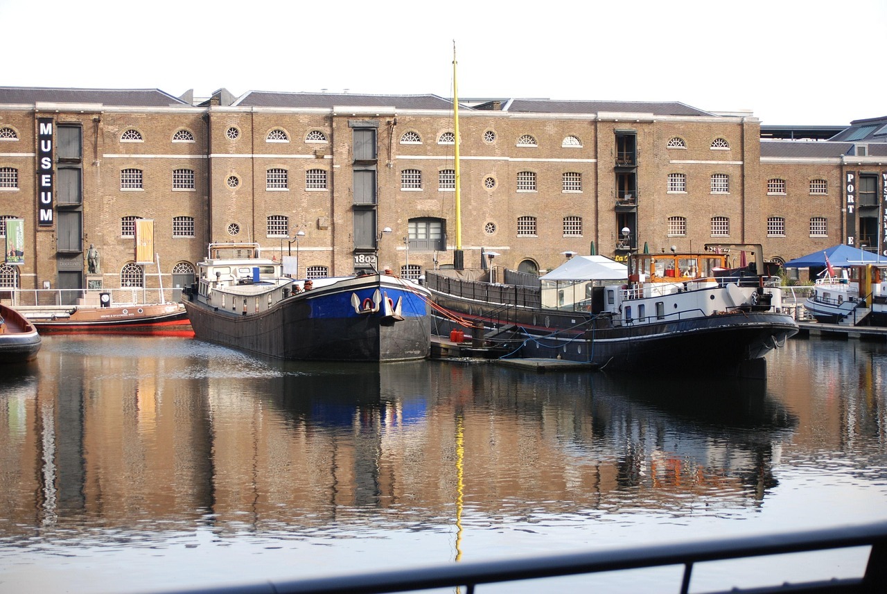 Two moored boats sit in calm water in front of the Museum of London Docklands, a historic brick warehouse building with arched windows. The scene includes reflections in the water and additional boats and signage in the background.